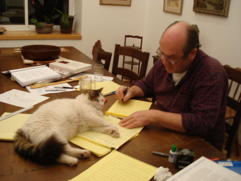 איציק מתפרס על שולחן חדר האוכל בבית כשהחתול ז'וז'י שומר עליו בעת כתיבת 'יום השנה לקארל שיינפלד'